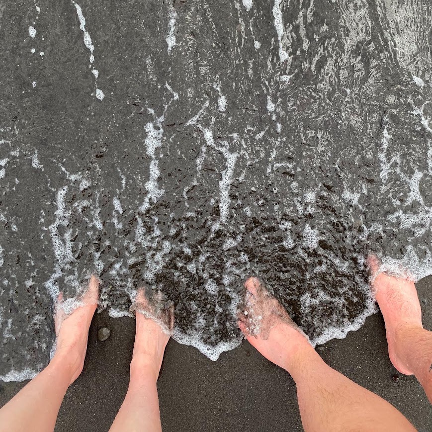 Two people's feet in black sand being washed over by a wave.