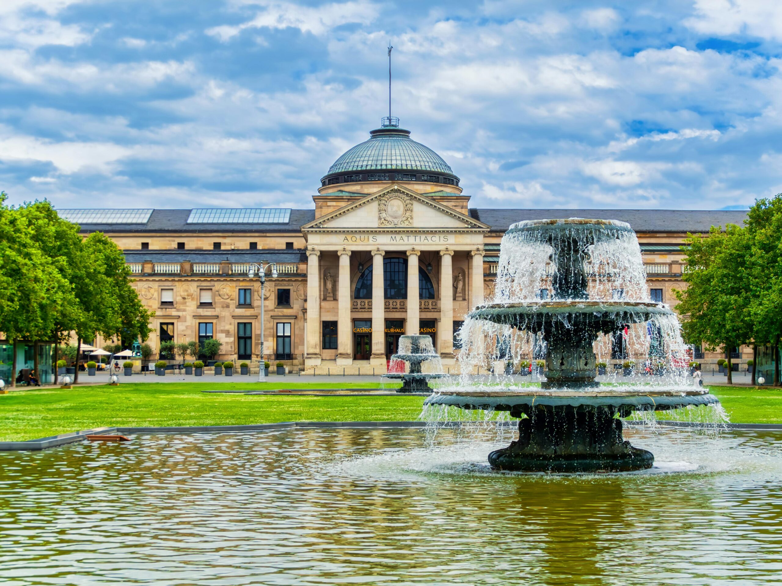 Wiebaden, Germany's Kurhaus with a fountain in the foreground under a bright sky with puffy white clouds.