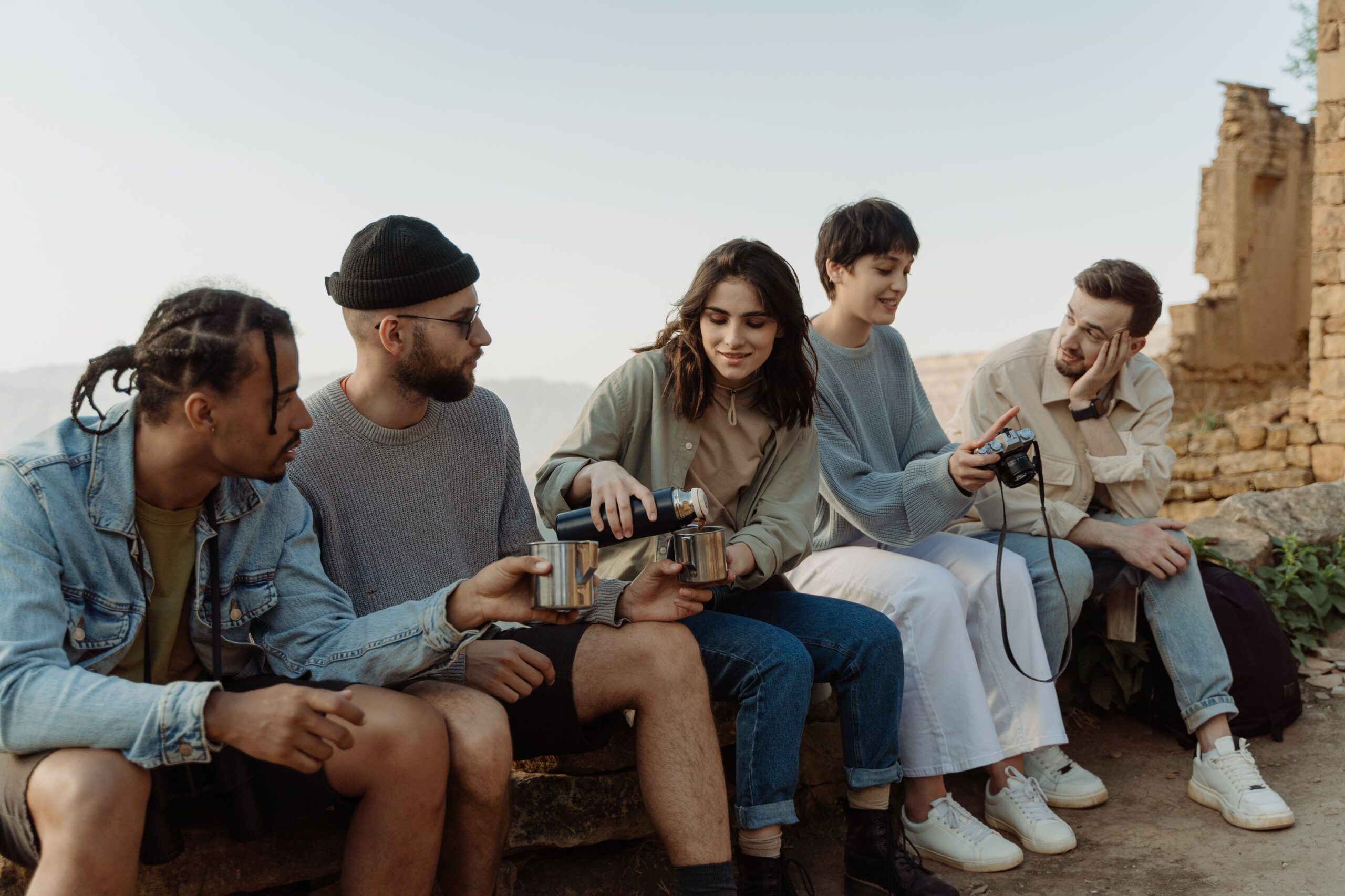 A diverse group of friends enjoy hiking together, sitting and sharing drinks outdoors on a sunny day, possibly on a group trip.
